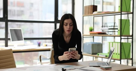 Female manager types message on mobile phone leaning on table in company premise. Brunette employee takes break enjoying communication with partner slow motion