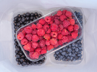 Three plastic containers with raspberries, blueberries and black currant in a white paper bag. Seasonal food shopping and healthy eating concept.