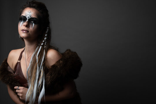 Studio portrait of a caucasian woman dressed up as a viking. She is wearing tribal makeup and has her hair in braids. She is wearing a bra and fur. There is empty space for text. 