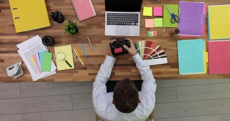 Professional photographer checks taken photos holding camera in hands. Man sits at big wooden table near open laptop and color palette fan upper view