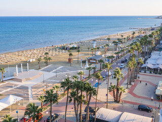 Top aerial view overlooking Finikoudes promenade with palm trees near the central sandy beach with sunbeds and sun glistening on blue water on a sunny day in Larnaca old town, Cyprus.