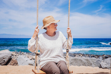 Happy senior woman sitting on swing on sea beach enjoying relaxed and carefree moments, positive...