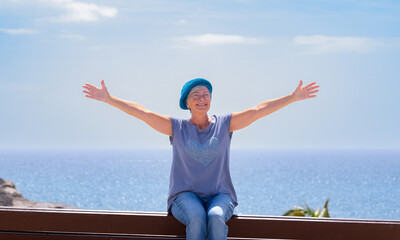 Cheerful attractive senior woman with open arms sitting outdoors on a bench at sea. Elderly lady relaxing enjoying vacation or retirement under the sun.  Horizon over water