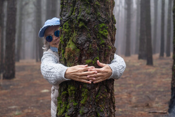 Blurred smiling senior woman with cap and blue glasses hugging a tree trunk in mountain forest on a...