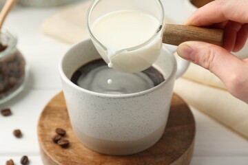 Woman pouring milk into cup with coffee at white table, closeup