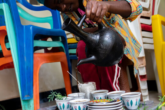 A jebena sits on a grass-covered table with small, handle-less cups arranged around it. Roasted coffee beans, a mortar and pestle, and incense smoke create a traditional Ethiopian coffee ceremony