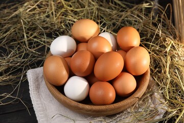 Fresh chicken eggs in bowl and dried hay on black wooden table