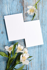 Two blank cards in portrait orientation placed on a blue wooden surface, accompanied by a cluster of white Alstroemeria flowers