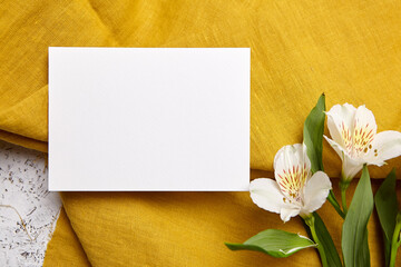Top view of a landscape-oriented white card on mustard-colored fabric, complemented by white Alstroemeria flowers
