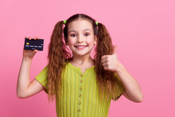 Photo portrait of cute little girl hold credit card thumb up dressed stylish green clothes isolated on pink color background