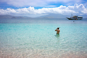 A man wades into the pristine waters off a pink sand beach, with a scenic boat and distant...
