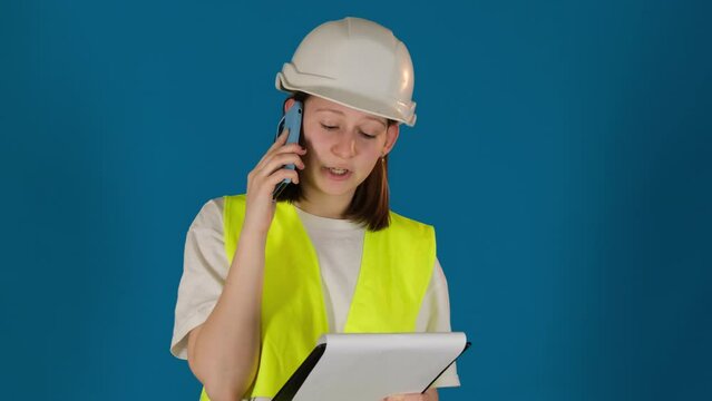 Female taskmaster in hardhat gives instructions via cellphone on blue background. Construction site inspector with clipboard reports boss by call