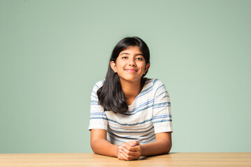 Indian asian small girl sitting at table against plain green background