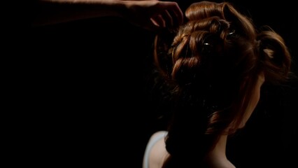 Hairdresser styling a young woman's reddish-brown hair in an intricate braid, with a close-up of the back of her head, highlighting the hair texture against a dark background.