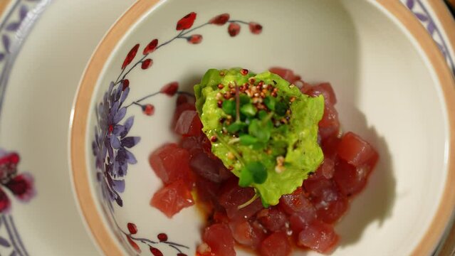 Close up of fresh tuna tartare with guacamole, micro greens and dijon mustard seeds. Gourmet raw fish fillet chopped in cubes and covered with avocado sauce in bowl with elegant ornaments.