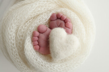 The tiny foot of a newborn baby. Soft feet of a new born in a white wool blanket. Close up of toes, heels and feet of a newborn. Knitted white heart in the legs of a baby. Macro photography