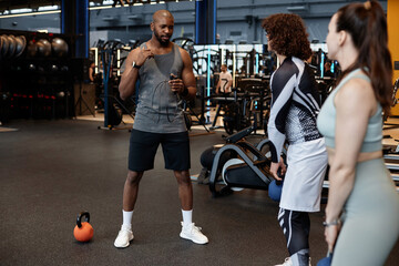 Full length portrait of muscular African American man as fitness coach talking to two people in gym...