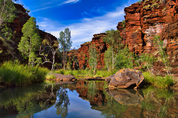 Red rocks, green trees and a huge boulder reflecting in the clear water of a pool in the Kalamina Gorge, Karijini National Park, Western Australia
 - Powered by Adobe