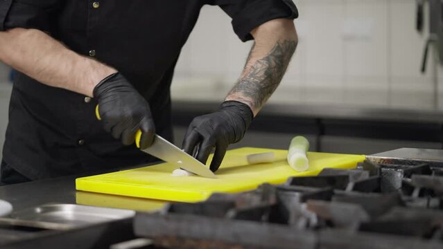 Close-up a professional chef in a black uniform and rubber gloves slices leeks on a yellow cutting board in a restaurant kitchen. Experienced chef expertly cuts vegetables while preparing a dish in