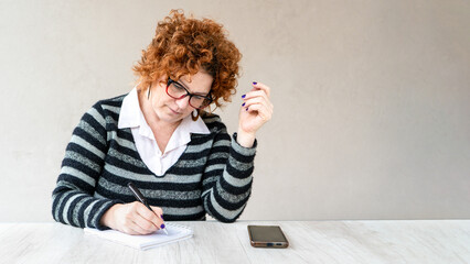 A woman with red hair and glasses is writing in a notebook. She has a cell phone on the table in...