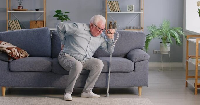 Senior man with walking stick is suffering from backache, lumbar ache or spine pain at home. Discomfort and challenges faced by seniors dealing with back injuries or spine related diseases.