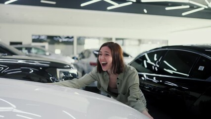 A happy and joyful brunette girl in a green jacket cheerfully hugs her new white car and dances and jumps near it after purchasing it at a car showroom