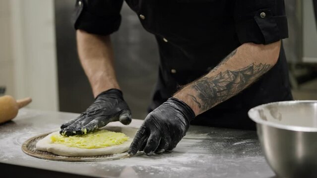 Close up a cook spreads a mixture of cheese onto a prepared flatbread of dough for making a cheese flatbread on a table on which there is flour and kitchen utensils for baking in a restaurant