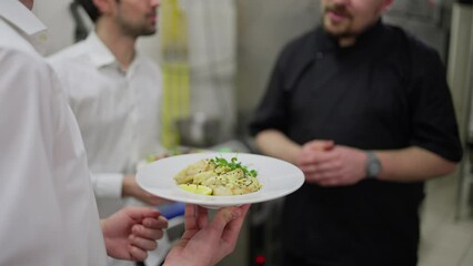 Close up a waiter in a white shirt holds a ready dish in his hands and communicates with a professional chef in a black uniform about serving it together with his waiter colleague in the kitchen in a