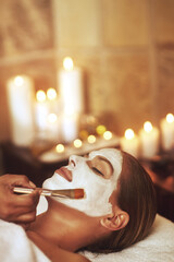 Woman, face mask and hand brush in spa for holiday treatment or beautician, stress relief or dermatology. Female person, calm and facial with candle bokeh at Bali resort in hotel, relax or skincare