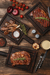 Wooden table served with various grilled meat, vegetables and glasses of beer. Striploin steak, ribeye steak and lamb ribs on wooden cutting boards. Top view