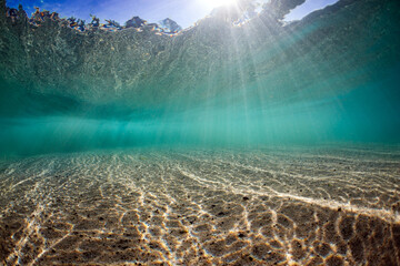 Shallow ocean with textured beach sand and aqua water filled with piercing light from above