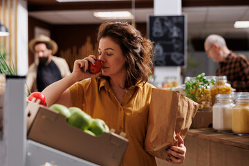 Woman smelling organic farm grown apples in busy zero waste store with low carbon emissions. Client...