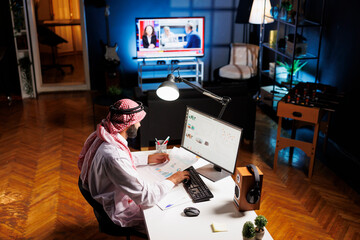 Muslim guy with paperwork diligently conducting online tasks efficiently. Young man in Islamic...