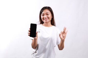 Portrait of a lovely young asian woman holding mobile phone and showing ok gesture isolated over white background