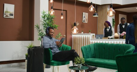 Tourist listens to music in lounge area, waiting to register at front desk and start international vacation. African american customer using headset and tablet to pass time, having fun.