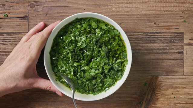 Cook mixes oil with chopped green fresh parsley in white bowl to make a healthy greens dressing. Close-up of food on kitchen wooden table background, top view