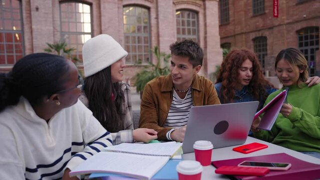 Group of diverse students talking animated working together using at laptop outdoor. Studying for exams and sharing knowledge sitting at table on university campus. Education technology gen z people