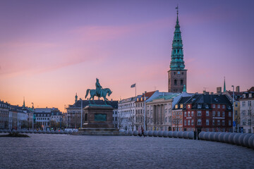A statue of a man riding a horse stands in front of a large building. The sky is a beautiful mix of pink and purple hues, creating a serene and peaceful atmosphere. Copenhagen - Powered by Adobe