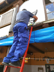 bricklayer construction worker on metal staircase to repair old tile roof and  and pvc gutters to collect rainwater