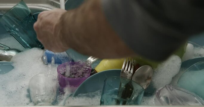 A man is washing dishes in a kitchen sink, scrubbing utensils and plates with soap and running water.