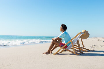 At beach, young biracial man sitting on beach chair, looking at sea