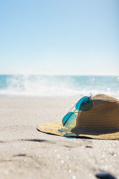 Sun hat and sunglasses on sandy beach; waves crash against a blue backdrop