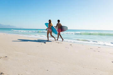 At beach, diverse couple holding surfboards, walking on sand