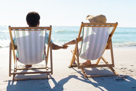 At beach, diverse couple holding hands, sitting on beach chairs, gazing at sea