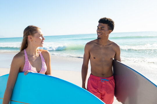 At beach, diverse couple holding surfboards, laughing together - Powered by Adobe