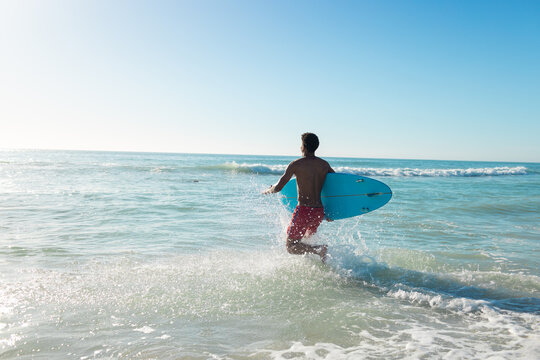 At beach, young biracial man running into sea holding surfboard
