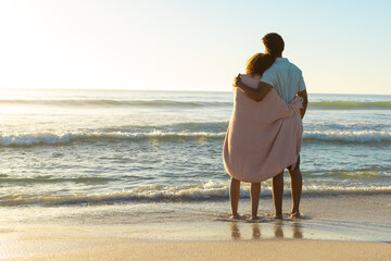 Obraz premium At beach, diverse couple standing, wrapped in blanket, gazing at sea