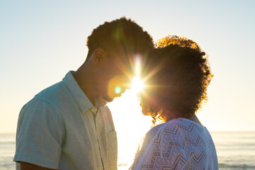 Obraz premium At beach, biracial couple standing close, sunset casting warm light on their faces