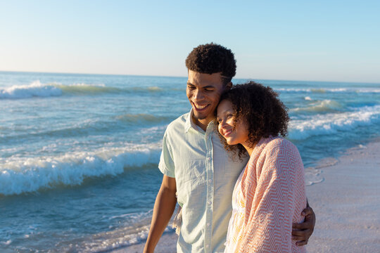 At beach, diverse couple embracing, walking on sand