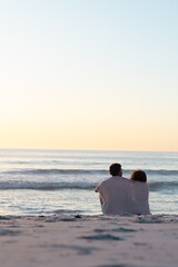Obraz premium At beach, biracial couple sitting, wrapped in blanket, looking at sea